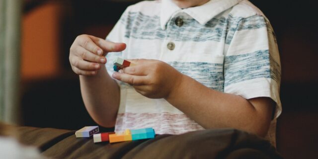 Kleinkind setzt LEGO-Bauteile zusammen, man sieht seinen Oberkörper und sein Gesicht bis zur Nase