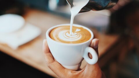 Person schüttet Milch aus einer Kanne in eine Tasse mit Kaffee und gestaltet Latte Art