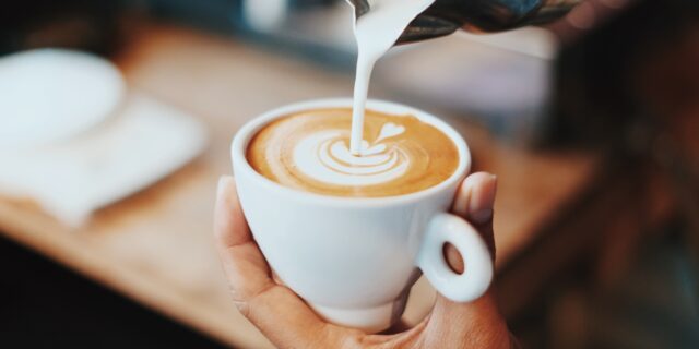 Person schüttet Milch aus einer Kanne in eine Tasse mit Kaffee und gestaltet Latte Art