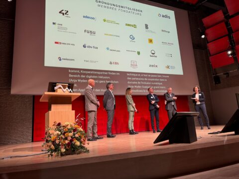 Sechs elegant gekleidete Personen auf einer Bühne, dahinter eine Leinwand mit den Logos der ADIS-Gründungsmitglieder