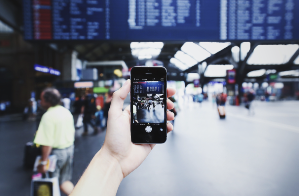 Eine Hand hält ein Smartphone in einer Bahnhofshalle. Auf dem Display ist die Anzeigetafel mit den Abfahrtszeiten zu sehen, die im Hintergrund unscharf über der Halle hängt.