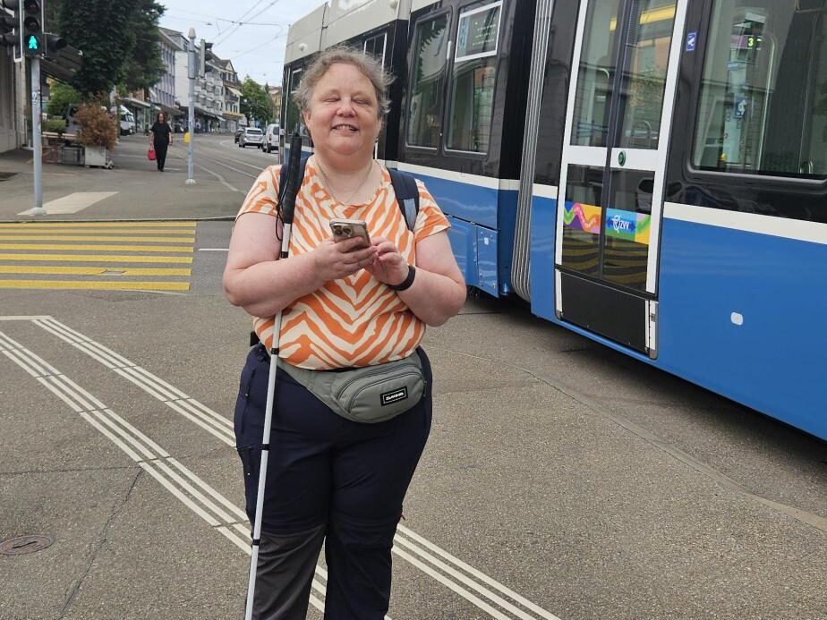 Regina se tient sur le trottoir, le sourire aux lèvres, son smartphone à la main, tandis qu'un tram passe devant elle.
